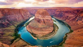 A view at sunset looking down at the Horseshoe Bend in the Colorado River near Page, Arizona. The river coils far below the viewer and is surrounded by colorful stone formations.