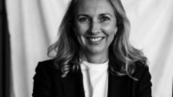 Black and white headshot of a woman with long blonde hair smiling into the camera, wearing a business blazer over a white T shirt, her arms crossed