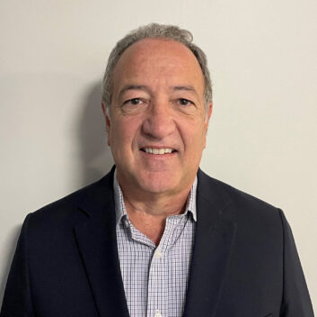 A man smiles at the camera. He wears a dark blazer over an open-necked business shirt.