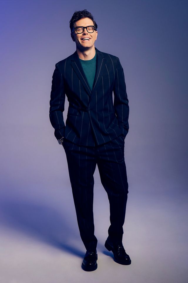 A man stands smiling with his hands in his pockets in a photo studio. He's wering a dark blue suit with big pinstripes over a blue T-shirt.