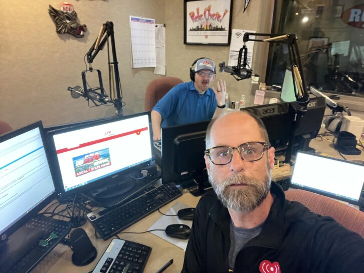 Two men sit in a radio studio, looking at the camera