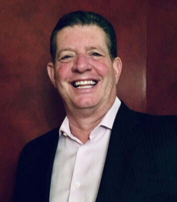 A man smiles at the camera. He wears a white open-necked business shirt and a dark blazer.