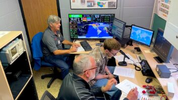 Three people sit at a table in a small windowless room. The young man in center is looking down at notes on the tabletop. The older men on either side of him are helping him learn about the amateur radio equipment in front of him including a transceiver, computer monitors and a screen showing a map of the world.