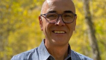 A man stands smiling at the camera, he is outside under some trees. He wears black-rimmed glasses and has an open-necked business shirt.