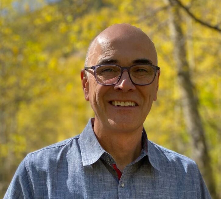 A man is smiling at the camera, standing outside under some trees. He wears black-rimmed glasses and an open-necked business shirt.