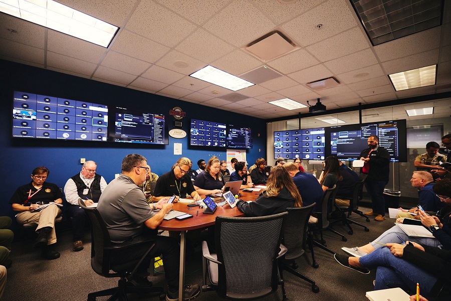 A room with many computer and video monitors on the walls, with a group of people sitting around a table in the center of the room