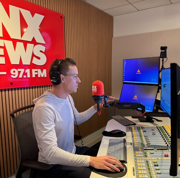 A man sits at a microphone in a radio studio, with a sign behind him that says "KNX News 97.1 FM"