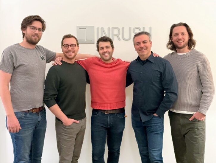 Five men stand smiling in front of a wall that bearns the logo of Inrush Broadcast Services