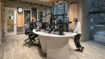 A bright newly constructed radio studio. A person sits at left, another stands at right at a guest position.